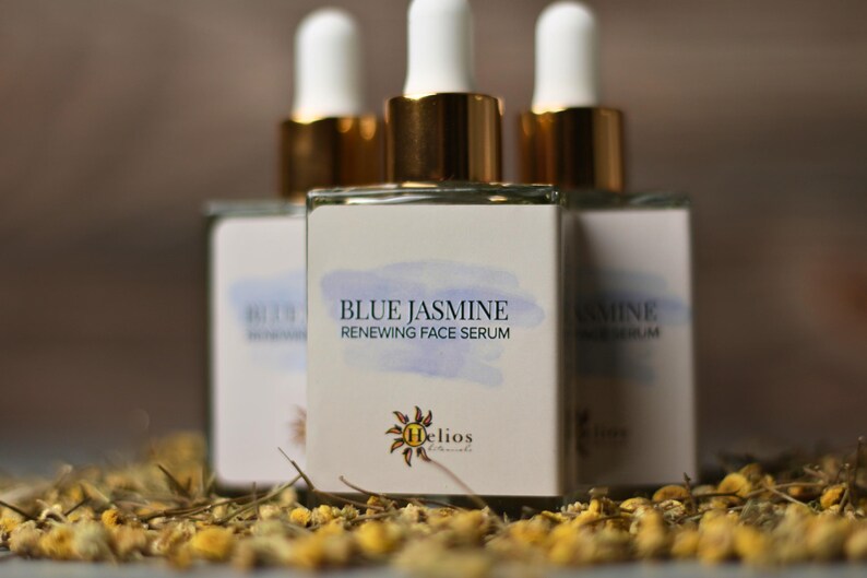 Op de afbeelding: Drie glazen flessen Blue Jasmine Renewing Face Serum met witte druppelaars en gouden accenten. De etiketten tonen de productnaam en een aquarelontwerp. De flessen zijn omgeven door gedroogde bloemen.