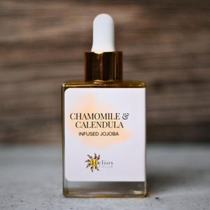May include: A square glass bottle with a white label featuring the text "Chamomile & Calendula Infused Jojoba." The bottle has a gold-colored top with a white dropper. The background is a blurred wooden surface.