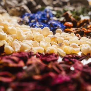 Op de afbeelding: Close-up van een verscheidenheid aan gedroogde botanische producten. De afbeelding toont lagen van ingredi&euml;nten, waaronder lichtgele hars, dieprode rozenblaadjes, blauwe bloemen en andere gedroogde kruiden. De focus ligt op de texturen en kleuren van de natuurlijke elementen.