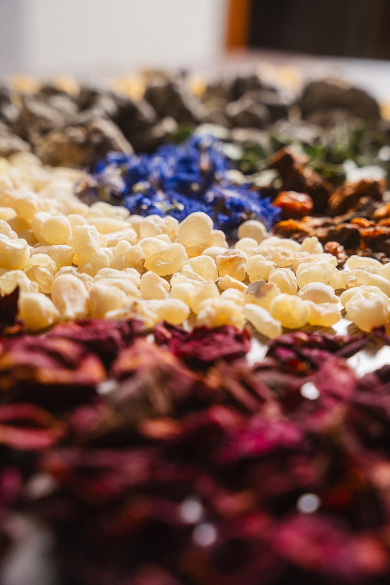Op de afbeelding: Close-up van een verscheidenheid aan gedroogde botanische producten. De afbeelding toont lagen van ingredi&euml;nten, waaronder lichtgele hars, dieprode rozenblaadjes, blauwe bloemen en andere gedroogde kruiden. De focus ligt op de texturen en kleuren van de natuurlijke elementen.
