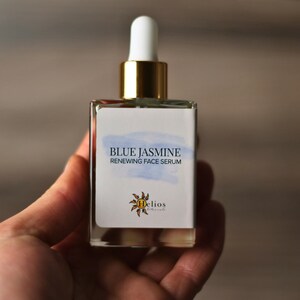 Op de afbeelding: Een heldere glazen fles Blue Jasmine Renewing Face Serum. De fles heeft een wit etiket met blauwe aquarelaccenten en gouden accenten op de dop. Het etiket bevat ook het Helios Naturals logo.