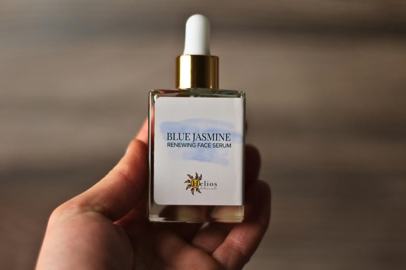 Op de afbeelding: Een heldere glazen fles Blue Jasmine Renewing Face Serum. De fles heeft een wit etiket met blauwe aquarelaccenten en gouden accenten op de dop. Het etiket bevat ook het Helios Naturals logo.