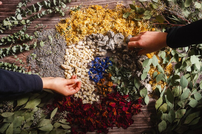 Op de afbeelding: Een cirkelvormige opstelling van natuurlijke elementen op een houten oppervlak. De compositie omvat gedroogde bloemen, stenen en kruiden in verschillende kleuren, waaronder geel, blauw, rood en groen. Twee handen zijn zichtbaar, die interageren met de opstelling.