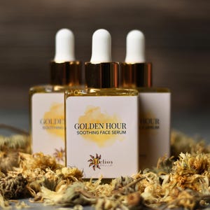 May include: Three bottles of Golden Hour Soothing Face Serum with white labels and gold accents. The labels feature the text "Golden Hour Soothing Face Serum" and a logo. The bottles are surrounded by dried flowers.