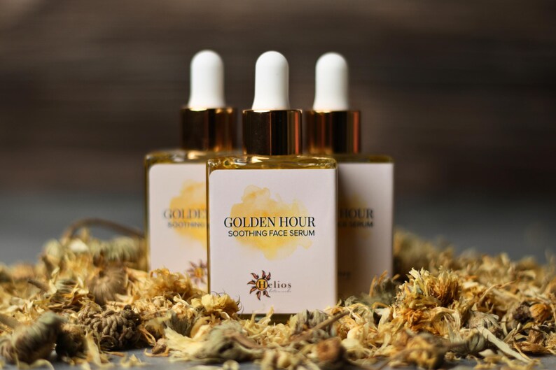 May include: Three bottles of Golden Hour Soothing Face Serum with white labels and gold accents. The labels feature the text "Golden Hour Soothing Face Serum" and a logo. The bottles are surrounded by dried flowers.