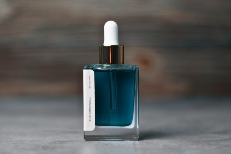 Op de afbeelding: Een heldere glazen fles gevuld met teal vloeistof, met een witte druppelaar en een goudkleurige kraag. Een wit etiket met zwarte tekst is op de fles bevestigd. De fles is gelabeld met "Helios Botanicals LLC" en "Burien WA".