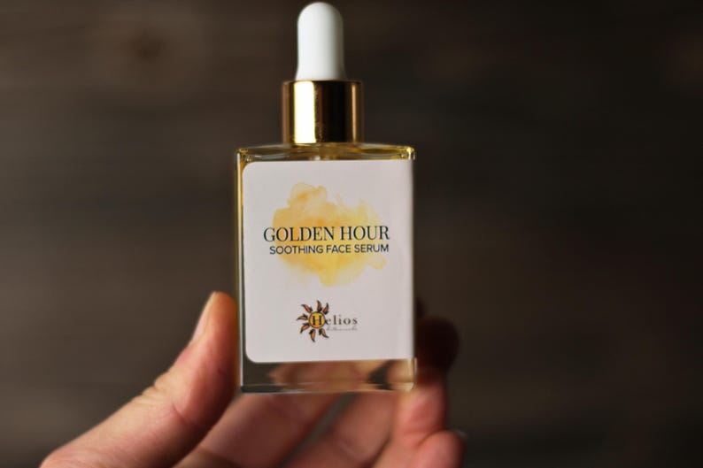 May include: A clear glass bottle of Golden Hour Soothing Face Serum. The bottle has a white label with gold text and a watercolor-style yellow design. The dropper is white and gold. The product is held in a hand.