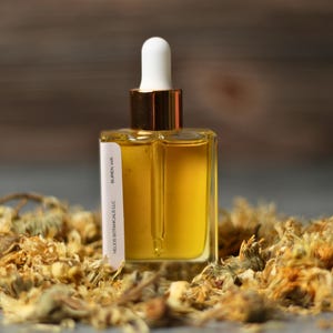 May include: A clear, square glass bottle with a white dropper top and gold accents, filled with a golden liquid. The bottle is labeled "HECO BOTANICALS LLC, BUREN WA" and is surrounded by dried flowers.