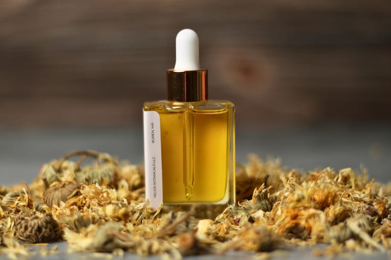May include: A clear, square glass bottle with a white dropper top and gold accents, filled with a golden liquid. The bottle is labeled "HECO BOTANICALS LLC, BUREN WA" and is surrounded by dried flowers.