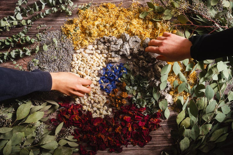 May include: A wooden surface displays an assortment of dried herbs and botanicals, including red rose petals, blue cornflowers, and yellow calendula. Hands are reaching into the arrangement. The scene suggests a natural, organic theme, possibly for crafting or aromatherapy.