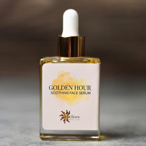 May include: A square glass bottle of Golden Hour Soothing Face Serum. The bottle has a white label with gold text and a yellow watercolor design. The dropper is white with a gold collar. The Helios Botanicals logo is at the bottom.