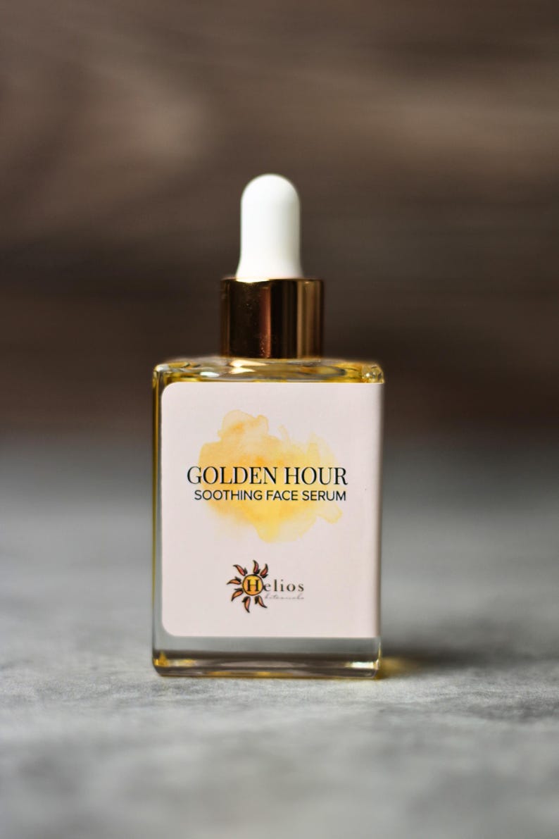 May include: A square glass bottle of Golden Hour Soothing Face Serum. The bottle has a white label with gold text and a yellow watercolor design. The dropper is white with a gold collar. The Helios Botanicals logo is at the bottom.