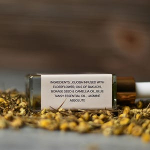 Op de afbeelding: Een kleine, rechthoekige glazen fles met een druppelaar, liggend op een bed van gedroogde gele bloemen. De fles heeft een gouden dop en een wit etiket met de ingredi&euml;nten: Jojoba, Vlierbloesem, Oli&euml;n van Bakuchi, Bernagiezaden & Camelia-olie, Blauwe boerenwormkruid essenti&euml;le olie, en Jasmijn Absolute.