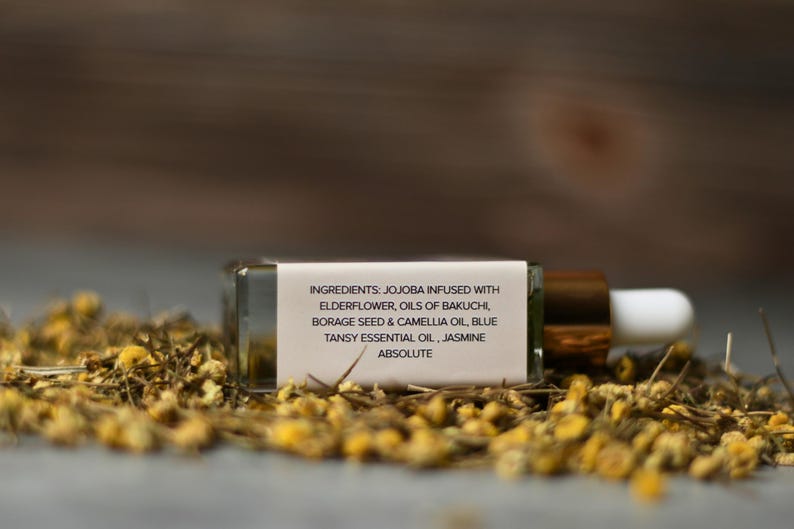 Op de afbeelding: Een kleine, rechthoekige glazen fles met een druppelaar, liggend op een bed van gedroogde gele bloemen. De fles heeft een gouden dop en een wit etiket met de ingredi&euml;nten: Jojoba, Vlierbloesem, Oli&euml;n van Bakuchi, Bernagiezaden & Camelia-olie, Blauwe boerenwormkruid essenti&euml;le olie, en Jasmijn Absolute.