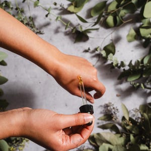 May include: A person's hands holding a dropper bottle, dispensing a golden liquid onto the skin. The background features green foliage and a light-colored surface, suggesting a natural skincare product.