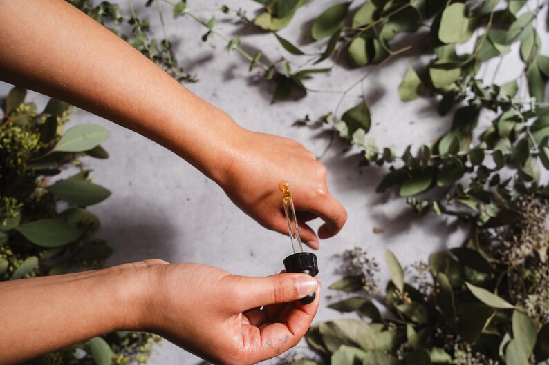 May include: A person's hands holding a dropper bottle, dispensing a golden liquid onto the skin. The background features green foliage and a light-colored surface, suggesting a natural skincare product.