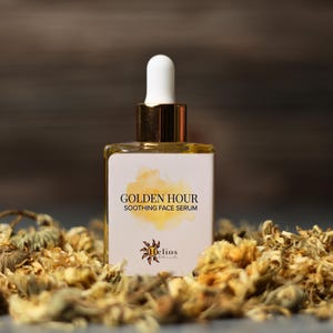 May include: A square glass bottle of Golden Hour Soothing Face Serum with a white dropper and gold accents. The label features the product name and a watercolor design. The bottle is surrounded by dried flowers, with a dark background.