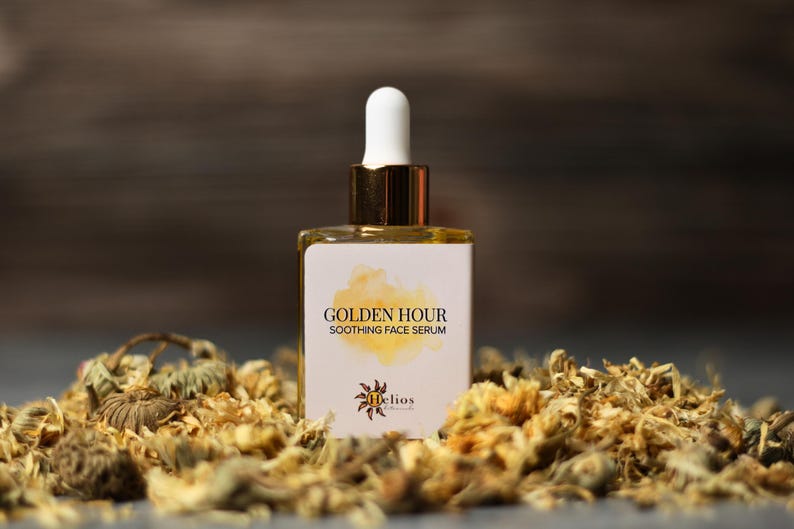 May include: A square glass bottle of Golden Hour Soothing Face Serum with a white dropper and gold accents. The label features the product name and a watercolor design. The bottle is surrounded by dried flowers, with a dark background.