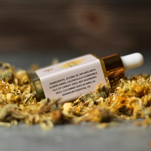 May include: A small glass dropper bottle with a gold cap and white dropper tip, resting on a bed of dried yellow flowers. The label displays the ingredients: jojoba oil, elderflower, calendula, chamomile, and other natural oils.