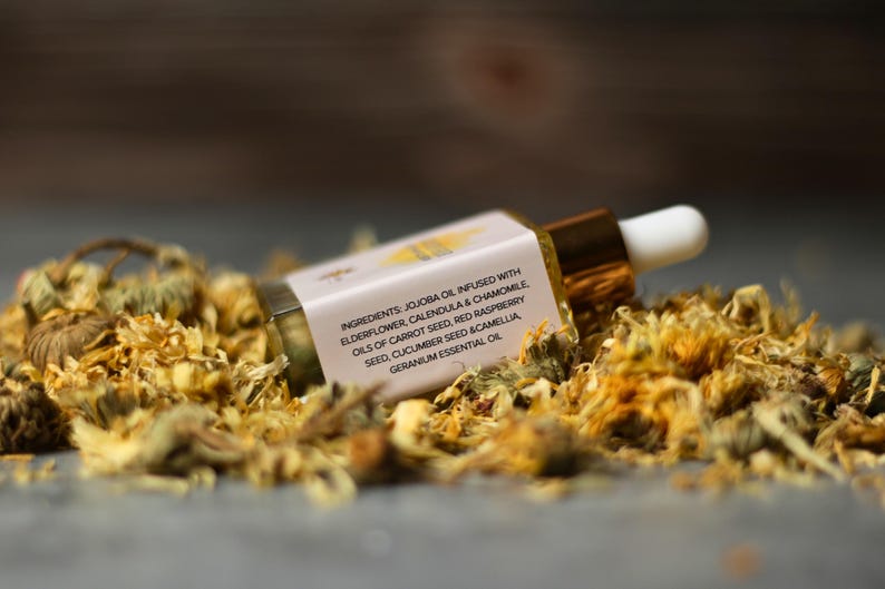 May include: A small glass dropper bottle with a gold cap and white dropper tip, resting on a bed of dried yellow flowers. The label displays the ingredients: jojoba oil, elderflower, calendula, chamomile, and other natural oils.