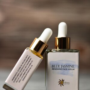 Op de afbeelding: Twee glazen flessen gezichtsserum. E&eacute;n fles leunt tegen de andere. De flessen hebben witte etiketten met de tekst "Blue Jasmine Renewing Face Serum" en informatie over de ingredi&euml;nten. De flessen hebben goudkleurige doppen en witte druppelaars.