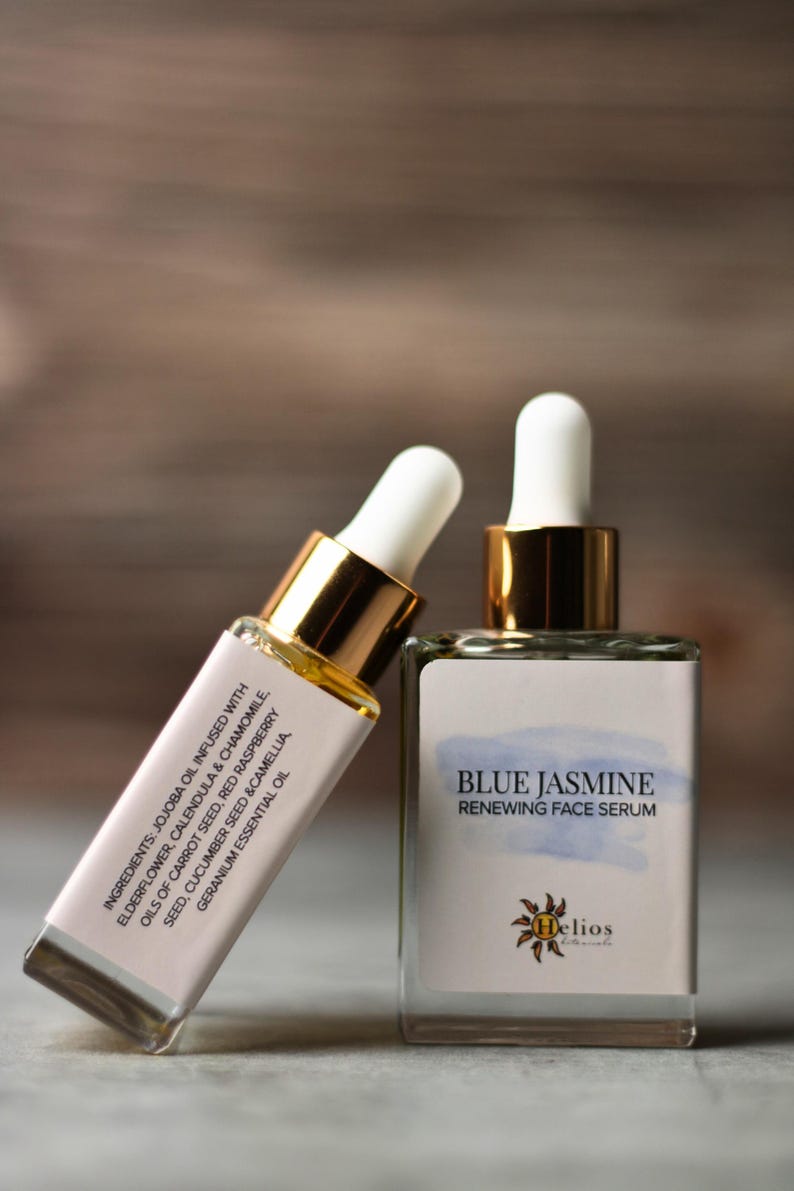 Op de afbeelding: Twee glazen flessen gezichtsserum. E&eacute;n fles leunt tegen de andere. De flessen hebben witte etiketten met de tekst "Blue Jasmine Renewing Face Serum" en informatie over de ingredi&euml;nten. De flessen hebben goudkleurige doppen en witte druppelaars.