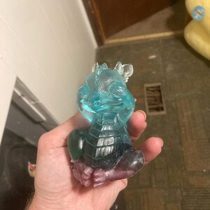 Könnte beinhalten: Eine durchscheinende Drachenfigur in Blau-, Grün- und Lilatönen. Der Drache sitzt und stützt seinen Kopf auf seine Hände. Die Figur hat einen strukturierten Körper und besteht aus einem transparenten Material.