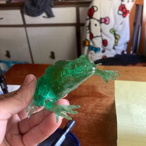 May include: A translucent green frog figurine with detailed features, held in a hand. The frog has visible legs and a textured body. The figurine is on a wooden table.
