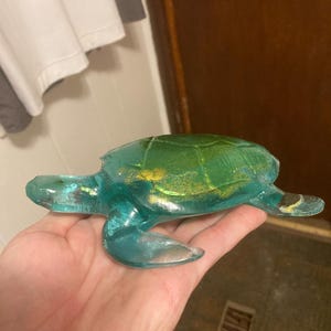 Puede incluir: Una figura de tortuga translúcida de color verde y turquesa con detalles de purpurina dorada. La tortuga se sostiene en una mano, mostrando su forma detallada y variaciones de color. La figura tiene un acabado liso y brillante.