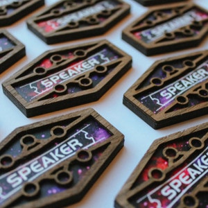 May include: Close-up of multiple wooden name tags with a galaxy print and the word "SPEAKER" in white lettering.
