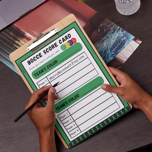 May include: A green and white printable bocce scorecard with the text "Bocce Score Card" and "Choose your team & write your name". The scorecard has a section for "Team's Color" with spaces for four players to write their names and colours. The scorecard is on a clipboard with a black pencil.