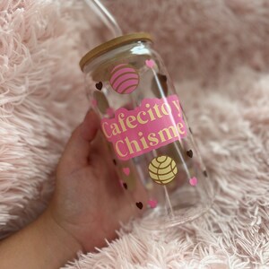 May include: A clear glass reusable water bottle with a pink and gold design. The bottle has a wooden lid and a straw. The design features the text "Cafecito y Chisme" and pink and yellow pastry designs.