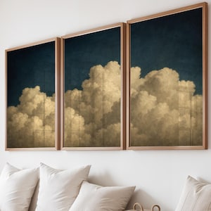 Può includere: Tre stampe artistiche incorniciate raffiguranti un paesaggio di nuvole con nuvole soffici e dorate su uno sfondo blu scuro. Le stampe sono in cornici di legno e sono appese sopra una panca di legno con cuscini bianchi e un cestino intrecciato.