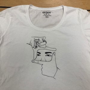 May include: White t-shirt with a black line drawing of two hands framing a face. The hands are making a frame with their fingers and the face is looking directly at the viewer.