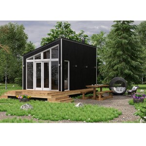 Op de afbeelding: Een klein zwart huisje met een groot raam en een houten terras. Het huisje is omringd door bomen en een grindpad.