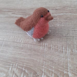 Könnte beinhalten: Eine kleine, handgefilzte Rotkehlchen-Figur aus brauner, grauer und roter Wolle. Der Vogel hat schwarze Augen und einen kleinen, spitzen Schnabel.