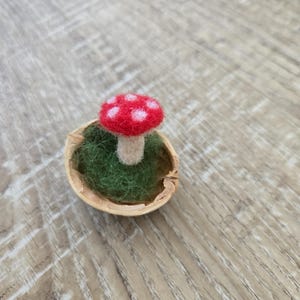 Könnte beinhalten: Ein Miniatur-Filzpilz mit rotem Hut und weißen Punkten sitzt auf grünem Filz in einer Walnussschale. Der Pilz hat einen beigen Stiel. Die Walnussschale liegt auf einer Holzoberfläche.