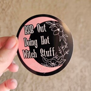 May include: A round sticker with a black crescent moon shape and pink border. The sticker has the text "BRB Out Doing Hot Witch Stuff" in white. Floral line art decorates the right side of the sticker.