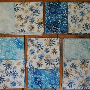 May include: A collection of folded fabric items with snowflake patterns in shades of blue, white, and gold. The fabrics are arranged on a wooden surface, showcasing a winter or holiday theme. The designs vary across the pieces.