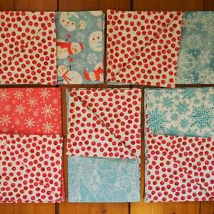 May include: An assortment of folded fabric squares with festive winter patterns. Designs include red polka dots, snowmen, snowflakes, and geometric shapes in red, white, and blue. The fabrics are arranged on a wooden surface.