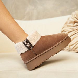 May include: Brown suede ankle boots with a faux fur lining and a decorative brown and white woven strap. The boots have a thick platform sole.