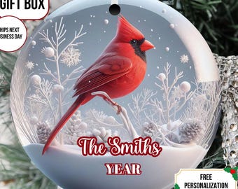 May include: A round Christmas ornament featuring a vibrant red cardinal perched on a snowy branch. The ornament has a winter scene with white accents and the text "The Smiths YEAR". Includes text that says "FREE GIFT BOX" and "FREE PERSONALIZATION".