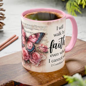 May include: A ceramic coffee mug with a pink interior and handle. The mug features a butterfly and floral design with the words "I will walk by faith even when I cannot see." The mug is filled with coffee.
