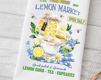 May include: White kitchen towel with a watercolor design featuring a jar of lemon curd, a teacup, and flowers. The towel has text that reads "Lemon Market" and "Hand-picked & Homemade Lemon Curd Tea Cupcakes."