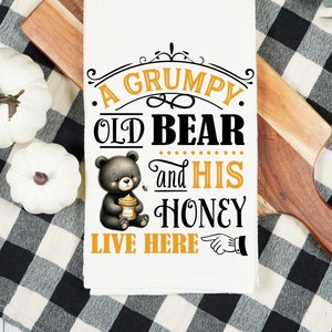May include: A white kitchen towel with a black and yellow design. The text reads "A Grumpy Old Bear and His Honey Live Here" with a cartoon bear holding a jar of honey.