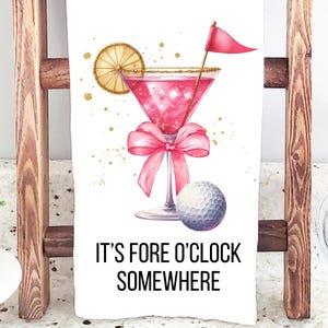 May include: White kitchen towel with a watercolor illustration of a pink cocktail with a golf flag, a golf ball, and a pink bow. The text "IT'S FORE O'CLOCK SOMEWHERE" is printed below the image. The towel is displayed on a wooden ladder.