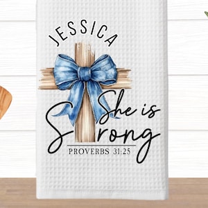 May include: White kitchen towel with a blue bow tied around a wooden cross. The text "She is strong" and "Proverbs 31:25" are printed below the cross. The name "Jessica" is printed above the cross.