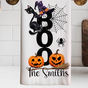 Op de afbeelding: Witte Halloween-keukendoek met een zwarte kat, heksenhoed, spinnenweb en twee pompoenen. Het woord "BOO" staat prominent afgebeeld, samen met "The Smiths" in een decoratief lettertype. Een bezem is ook inbegrepen.