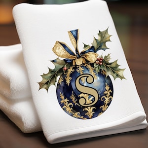 May include: White hand towel with a navy blue and gold Christmas ornament design. The ornament features the letter "S", gold filigree, a gold bow, and holly leaves. The towel is folded and stacked with two other white towels.