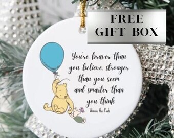 May include: A white, round Christmas ornament with a gold string, featuring a Winnie the Pooh illustration with a blue balloon. The ornament has a quote and the text "FREE GIFT BOX".